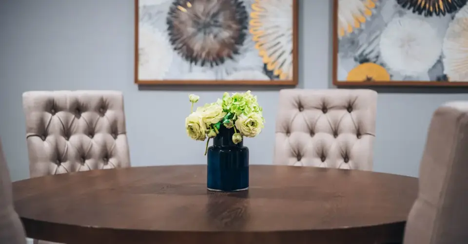 A round dining table, upholstered chairs, flowers, and framed wall art form a finished dining room.