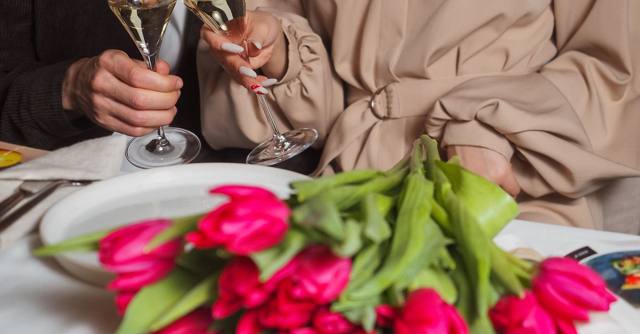 A couple raises champagne glasses above pink tulips at dinner.
