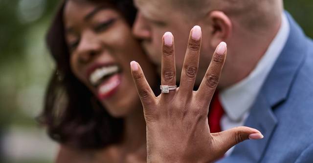 A newly engaged woman holds up her ring as her partner smiles behind her.