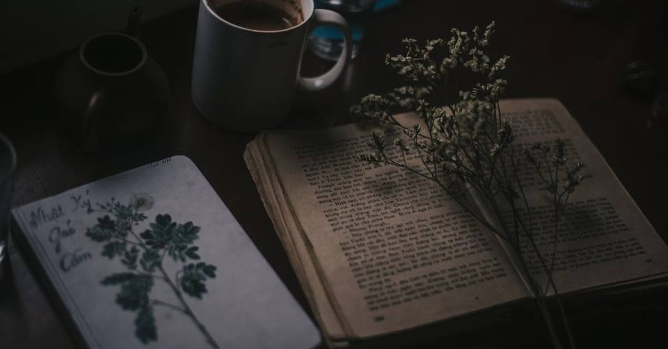 Moody still life of vintage book, coffee mug, and dried flowers on a dark wooden table.