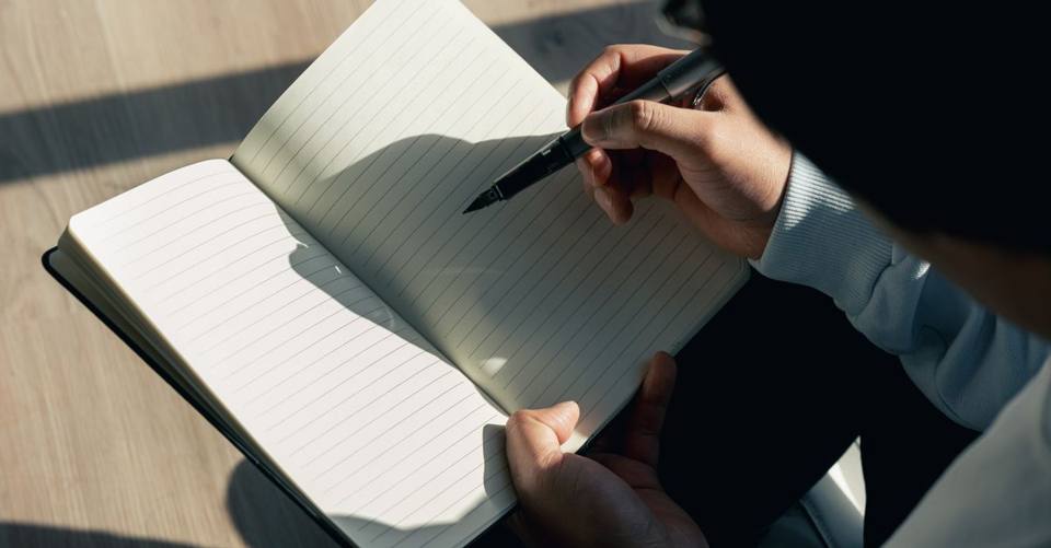 A person writes in a lined notebook in a patch of sunlight.