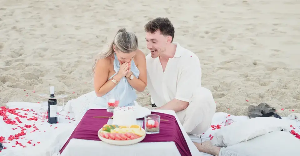 A couple enjoying a romantic picnic on the beach with a celebration cake and fruit.