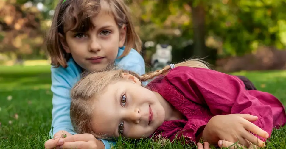 Two sisters enjoying a sunny afternoon, lying relaxed on the green grass in a peaceful park setting.