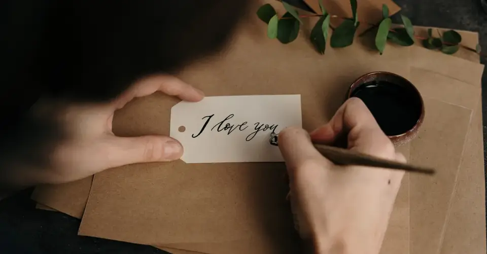 Hands use a dip pen to write “I love you” on a small card over a kraft-paper envelope.