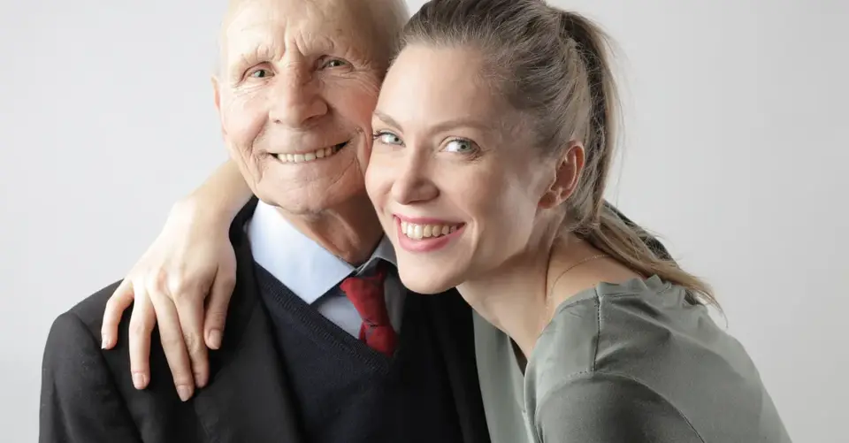 An older father and adult daughter embrace and smile at the camera.