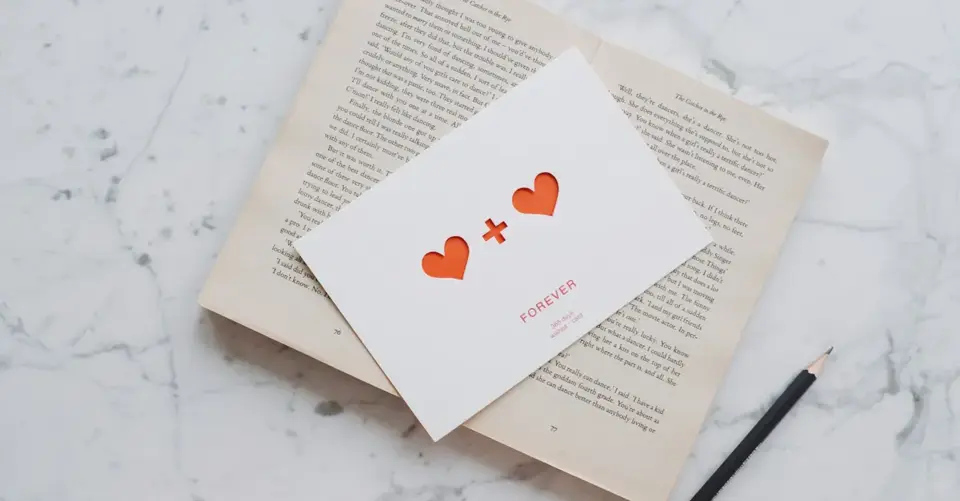A small card with orange hearts rests on an open book beside a black pencil.