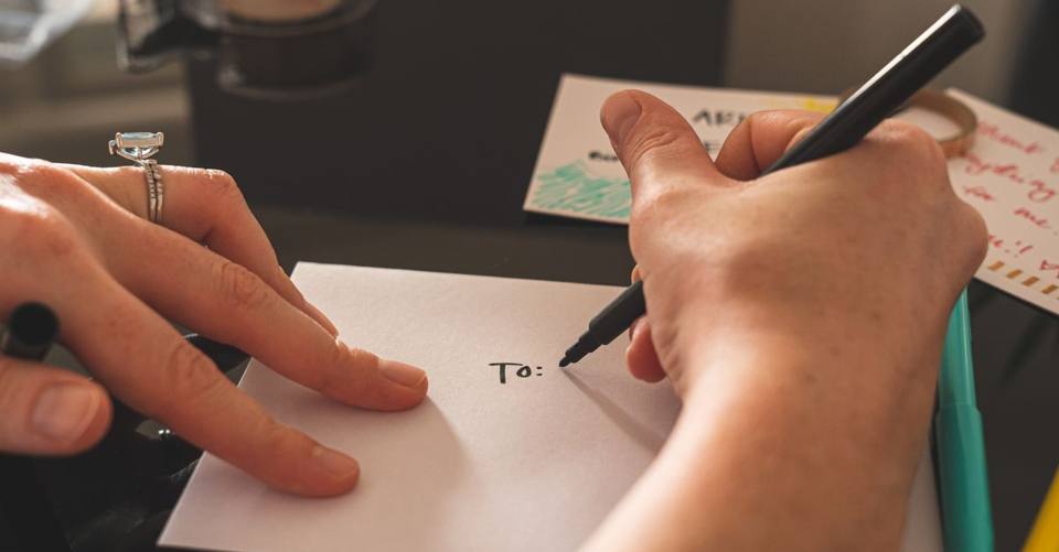 Hands write an address on a white envelope with a black marker.