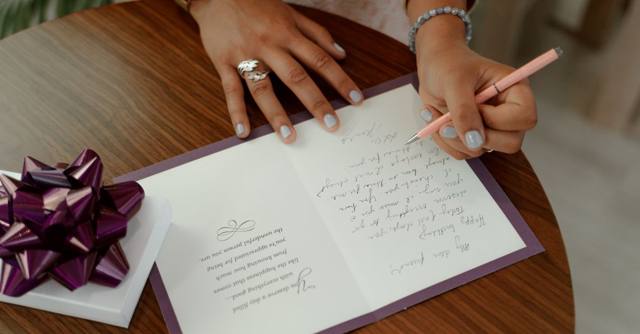 A woman writes a message in a greeting card beside a wrapped gift.