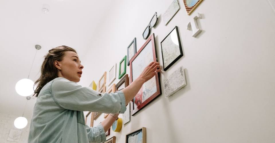 A woman adjusts a colorful gallery wall of framed artwork.