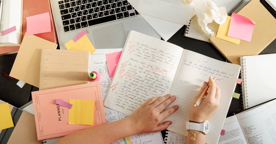 A person edits handwritten notes at a desk scattered with notebooks, sticky notes, and a laptop.