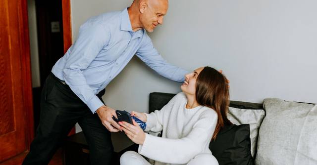 A father gives his adult daughter a gift as they sit together at home.
