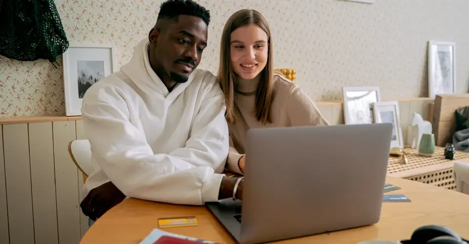 A smiling couple sit together at a table and review something on a laptop.