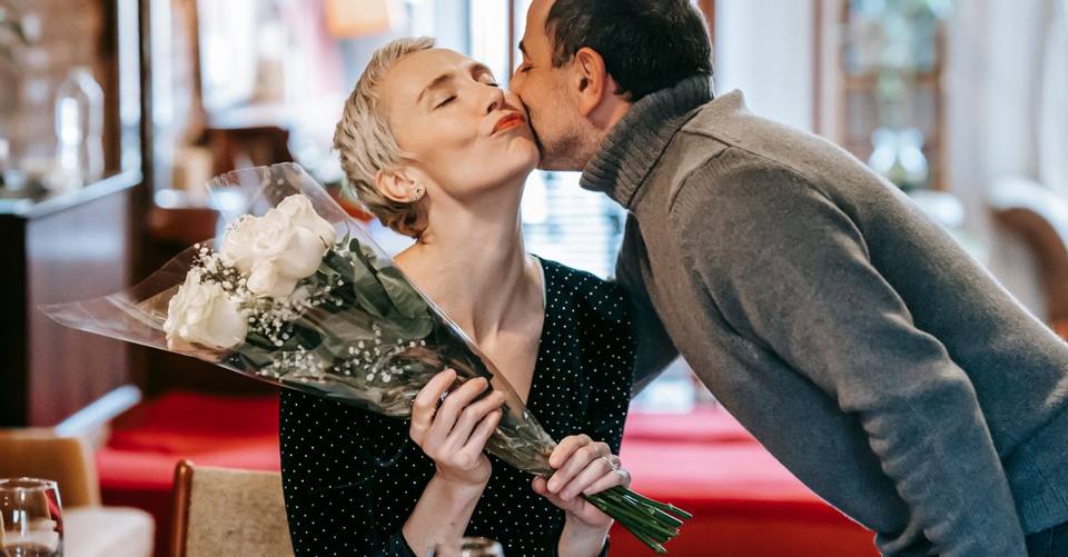Couple having romantic dinner while male kissing in cheek with closed eyes and giving bouquet with white roses to female near table with wineglasses and plate with pasta with meatballs