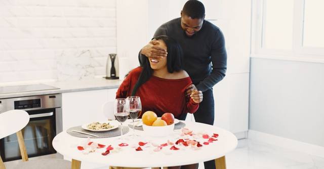 A man surprises his partner at a candlelit dinner table.
