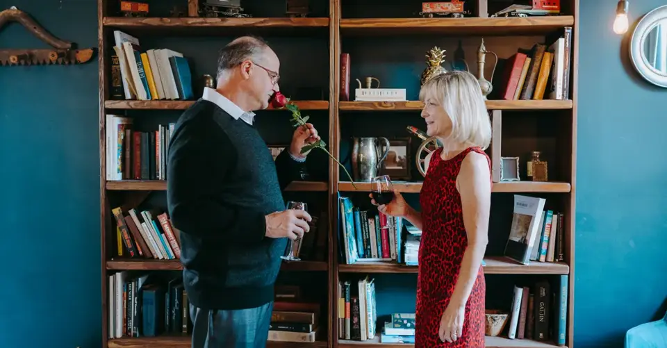 Elderly couple enjoying a romantic moment with rose and wine in a cozy home library.