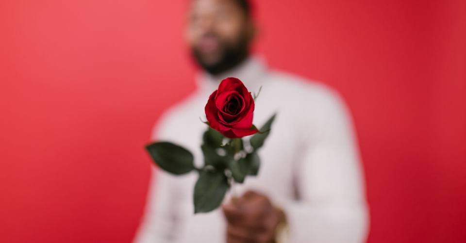 A man in blurry focus offering a red rose against a vibrant red background, symbolizing love.