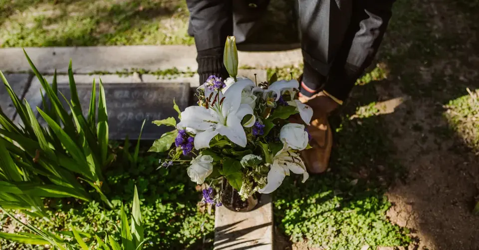 A man places a bouquet of flowers on a grave in a peaceful cemetery setting.