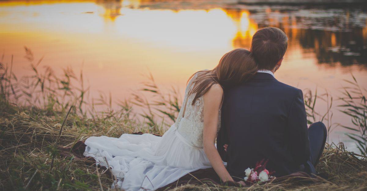 Newlywed couple sitting by a lake during sunset, embracing in a romantic setting.