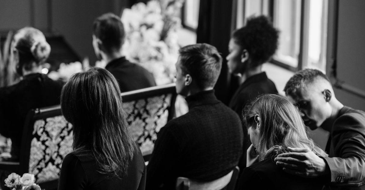 A somber grayscale image of mourners sitting indoors during a funeral, conveying loss and sympathy.