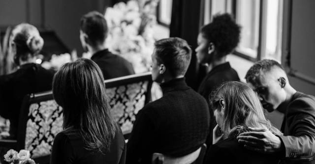 A somber grayscale image of mourners sitting indoors during a funeral, conveying loss and sympathy.