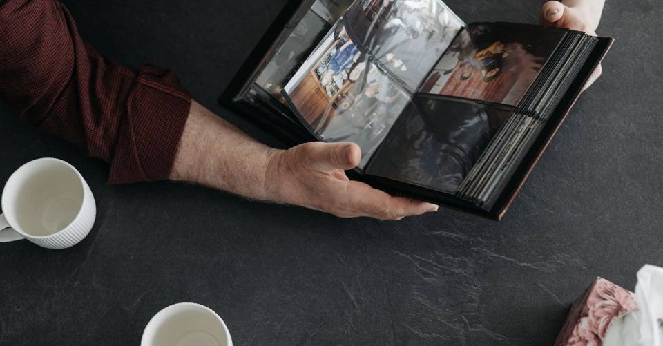 Hands hold open a photo album beside two coffee cups on a dark table.