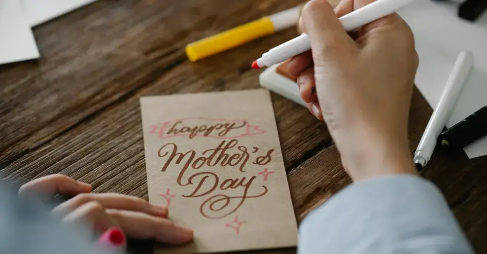 Hands letter a Mother’s Day card with brown ink at a wooden table.