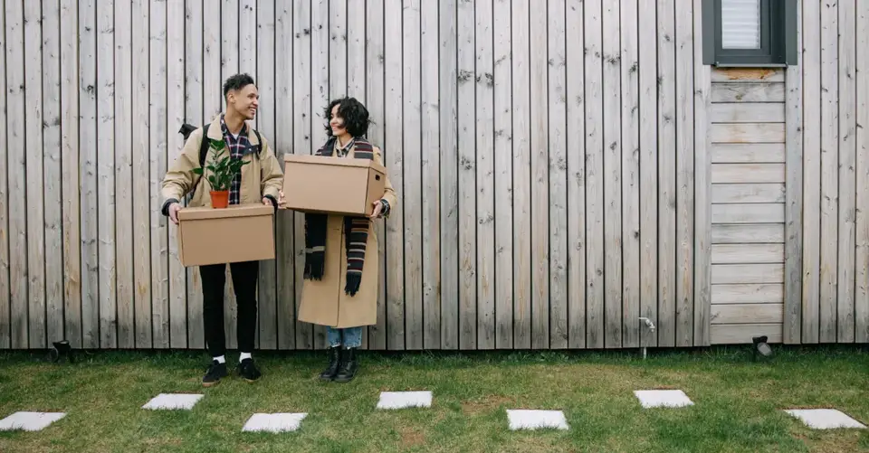 Happy couple carrying boxes and a plant, moving into a new home against a wooden wall.