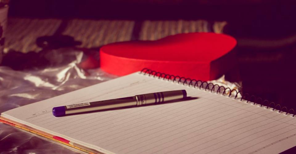 A spiral notebook with a pen placed on a heart-shaped box, symbolizing love and writing.