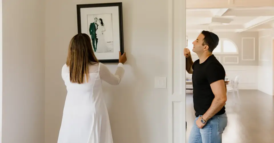 A couple align a framed photograph on a light-colored wall.